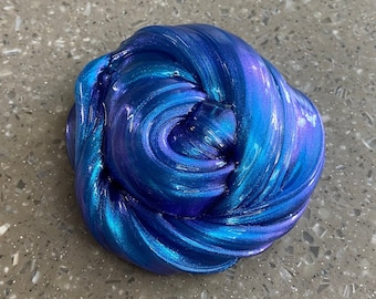 May include: A close-up of a blue and purple slime with a shimmery finish. The slime is folded and twisted into a ball shape.