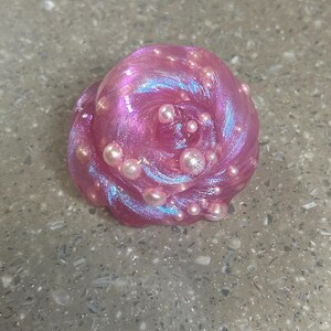 May include: A swirl of pink slime with iridescent highlights and small, pearl-like beads. The slime appears to be soft and pliable, with a glossy finish. The beads are scattered throughout the slime, adding texture and visual interest.