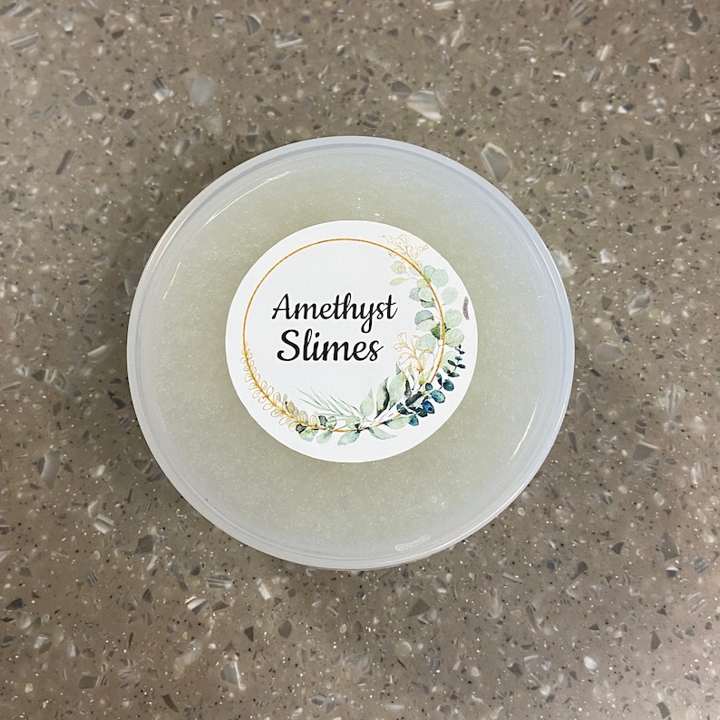 May include: A clear plastic container filled with a white slime with glitter. The container has a white label with a gold circle and green leaves around the text "Amethyst Slimes".