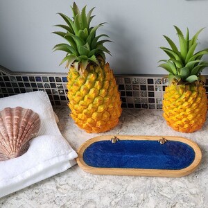 May include: Two yellow pineapple decorations and a white towel with a seashell on top. A blue and wooden tray with two gold turtle figurines sits on a gray countertop.