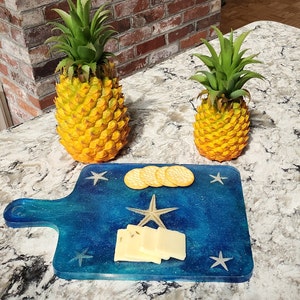 May include: A decorative serving board with a blue and turquoise epoxy resin design, adorned with starfish and a handle. The board holds crackers and cheese. Two yellow pineapples with green tops are also present.