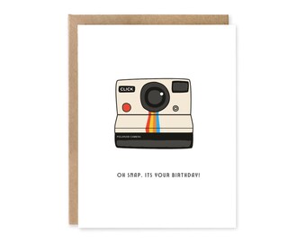 May include: A birthday greeting card featuring a cartoon illustration of a vintage Polaroid camera. The camera is off-white with a black lens and a colorful stripe. The text "OH SNAP. IT'S YOUR BIRTHDAY!" is below the camera.