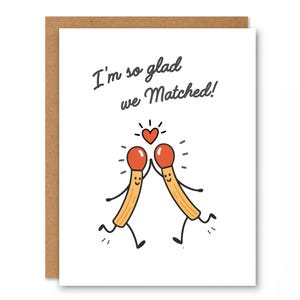 Puede incluir: Una tarjeta de felicitación con borde marrón e interior blanco. La tarjeta presenta el texto "I'm so glad we Matched!" encima de una ilustración de dibujos animados de dos cerillas con caras sonrientes, brazos y piernas, chocando los cinco bajo un corazón rojo.