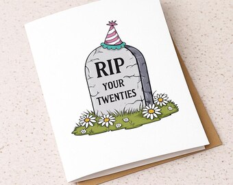 Puede incluir: Tarjeta de felicitación blanca con una lápida de dibujos animados que dice "RIP YOUR TWENTIES". La lápida tiene un gorro de fiesta encima y está rodeada de margaritas. La tarjeta está sobre una superficie clara.