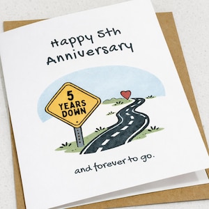 以下が含まれることがあります： 5周年記念のグリーティングカード。カードには「5 Years Down」と書かれた道路標識と、赤いハートへと続く曲がりくねった道が描かれています。「Happy 5th Anniversary」と「and forever to go」の文字も含まれています。