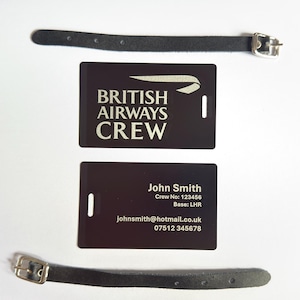 Op de afbeelding: Twee zwarte bagagelabels met de tekst "BRITISH AIRWAYS CREW" en persoonlijke gegevens. Elk label heeft een zwarte leren riem met een zilveren gesp. De labels zijn rechthoekig.