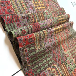 May include: A rolled-up textile with a patchwork design. The fabric features a mix of red, green, purple, and brown hues. The pattern includes floral and geometric motifs. The textile appears to be made of a soft, possibly silk, material.