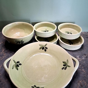 May include: A set of light green ceramic kitchenware featuring a hand-painted olive branch motif. The collection includes a large round bowl, a wide shallow dish with two side handles, and four smaller round bowls, all with a smooth glazed finish.