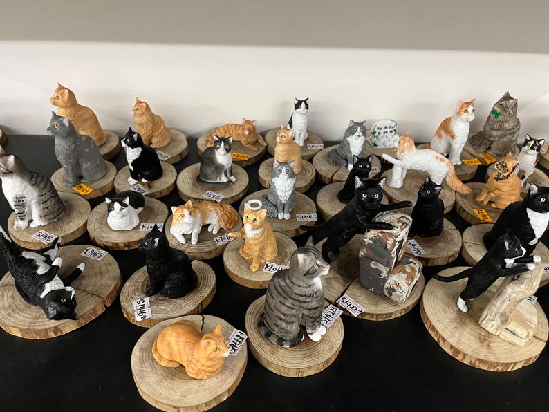 May include: A selection of cat ornaments in different colours and poses, such as orange, black, grey, and white, presented on small wooden slices. Some cats are sitting, standing, or playing. The ornaments are detailed and appear to be handmade.