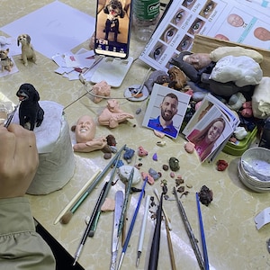 May include: A person is painting a small, black dog figurine with a paintbrush. The figurine is on a white pedestal. Other dog figurines, clay, tools, and reference photos are scattered on the table, suggesting a crafting or sculpting project.