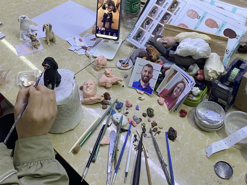 May include: A person is painting a small, black dog figurine with a paintbrush. The figurine is on a white pedestal. Other dog figurines, clay, tools, and reference photos are scattered on the table, suggesting a crafting or sculpting project.
