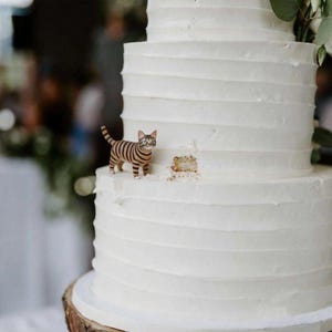 May include: A white wedding cake with multiple tiers and textured frosting. A small figurine of a tabby cat with brown and black stripes is placed on the cake. The cake is on a wooden base, with a small piece missing.
