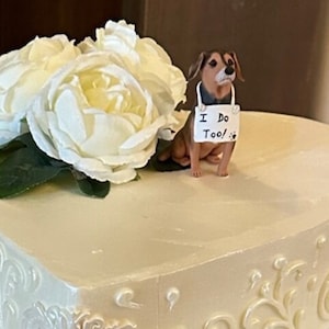 Może przedstawiać: Biały tort ślubny w kształcie serca ozdobiony białym lukrem i delikatnym wzorem kwiatowym. Na szczycie tortu znajduje się mała figurka brązowego i czarnego psa trzymająca tabliczkę z napisem "Ja też!"