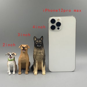 May include: Three dog figurines of varying sizes, approximately 5, 7.6, and 10 cm tall, are displayed next to an iPhone 12 Pro Max. The dogs are painted in realistic colours, with one wearing a green collar. The background is a solid grey.