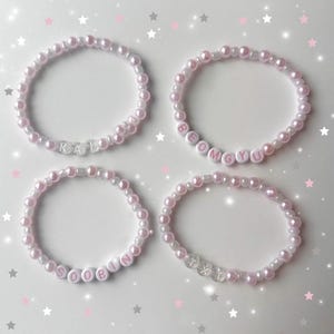 May include: Four beaded bracelets with pink and white pearls. Each bracelet has a different name spelled out in white letter beads: KAI, BEOMGYU, SOOBIN, and TXT. The bracelets are arranged on a white surface with pink and gray star decorations.
