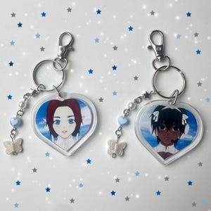 May include: Two heart-shaped keychains with anime-style character portraits. Each keychain has a silver clasp, a beaded chain with blue hearts and pearls, and a white butterfly charm. The background is a light blue sky.
