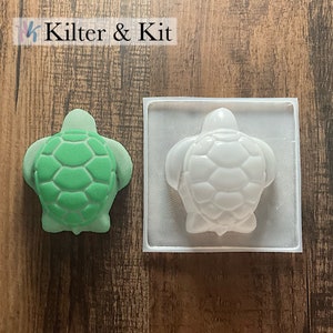 May include: A white plastic mold of a turtle, with a green turtle made of a similar material next to it. The mold is in a square frame.