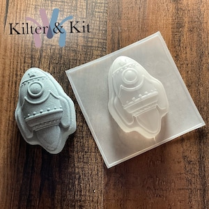 May include: A clear plastic mold of a rocket ship, with a gray rocket ship made from the mold next to it. The rocket ship has a round window, a pointed nose, and a fin on each side.
