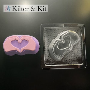 May include: A clear plastic mold for making soap or resin in the shape of two hands forming a heart. The mold is shown next to a purple soap bar with the same design.