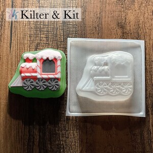 May include: A red and white train-shaped confection with gray wheels and a green base sits next to a clear plastic mold of the same design. The text "Kilter & Kit" is in the upper left corner.