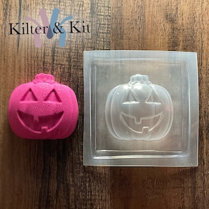 May include: A clear plastic mold of a pumpkin with a smiling face. A pink pumpkin with a smiling face is next to the mold.