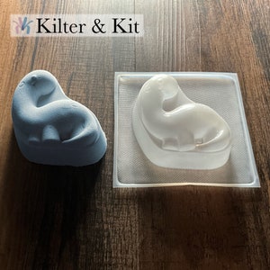 May include: A light blue dinosaur-shaped mold and a white dinosaur-shaped mold. The molds are made of silicone and are used for making soap, candles, or other crafts.