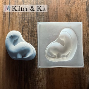 May include: A light blue dinosaur-shaped mold and a matching dinosaur made from the mold. The mold is made of clear plastic and has a textured surface. The dinosaur is made of a smooth, light blue material.