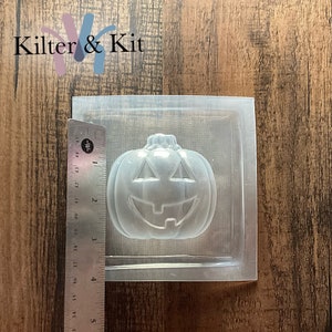 May include: Clear plastic mold for making a pumpkin-shaped soap or candle. The mold is approximately 3 inches wide and 3 inches tall. The mold has a detailed design of a pumpkin with a carved face.