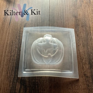 May include: Clear plastic mold for making a pumpkin-shaped soap or candle. The mold features a detailed jack-o'-lantern face with a wide smile and two eyes.