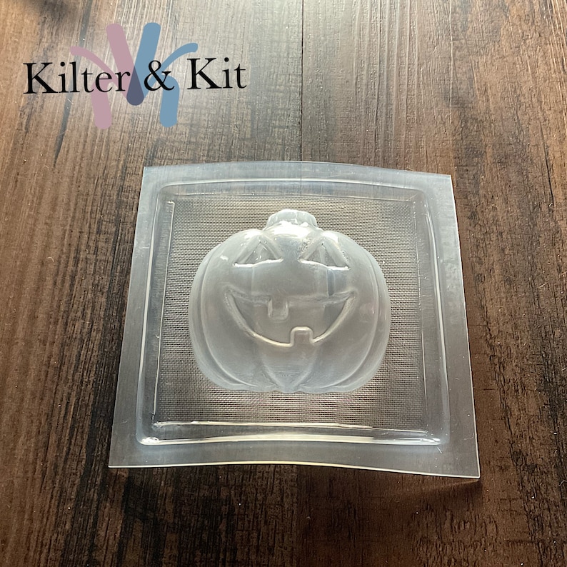May include: Clear plastic mold for making a pumpkin-shaped soap or candle. The mold features a detailed jack-o'-lantern face with a wide smile and two eyes.