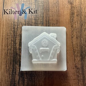 Puede incluir: Un molde de pl&aacute;stico transparente con forma de casa de jengibre con detalles de bast&oacute;n de caramelo. El molde est&aacute; sobre un fondo blanco.