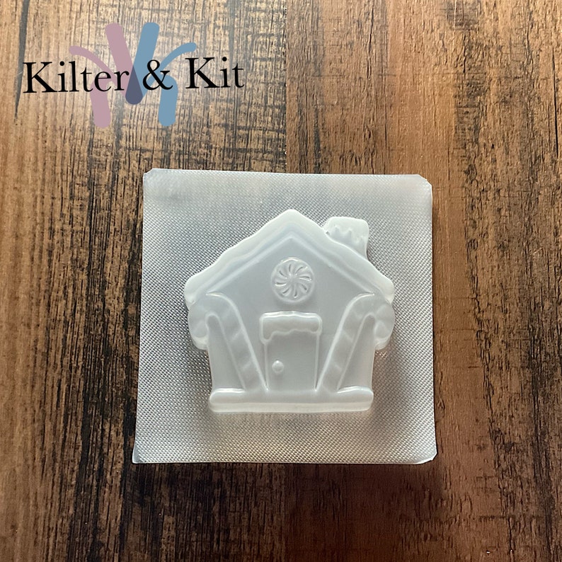 Puede incluir: Un molde de pl&aacute;stico transparente con forma de casa de jengibre con detalles de bast&oacute;n de caramelo. El molde est&aacute; sobre un fondo blanco.