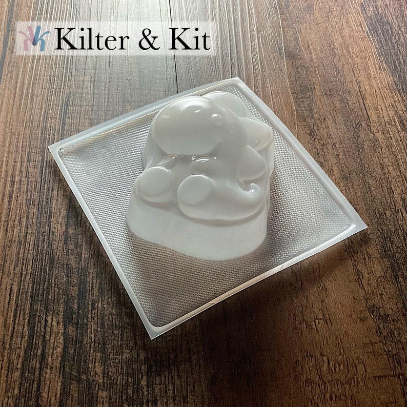 May include: Clear plastic mold for making a small elephant-shaped soap or candle. The mold is on a wooden surface.