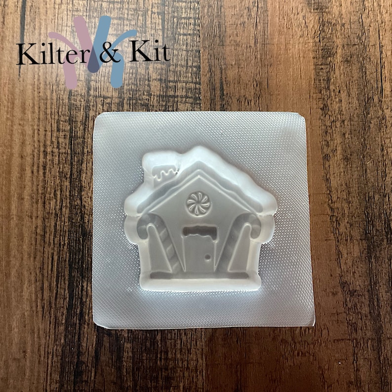 Puede incluir: Un molde de pl&aacute;stico transparente con forma de casa de jengibre con detalles de bast&oacute;n de caramelo. El molde est&aacute; sobre una superficie texturizada.