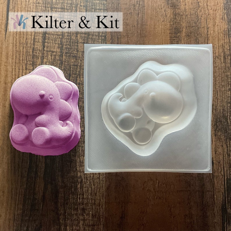 May include: A clear plastic mold of a cartoon dinosaur, with a purple dinosaur made from bath bomb mix in the mold.