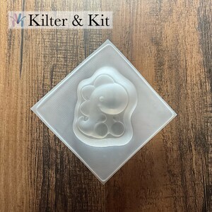 May include: Clear plastic mold shaped like a cartoon dinosaur. The mold is on a wood background.