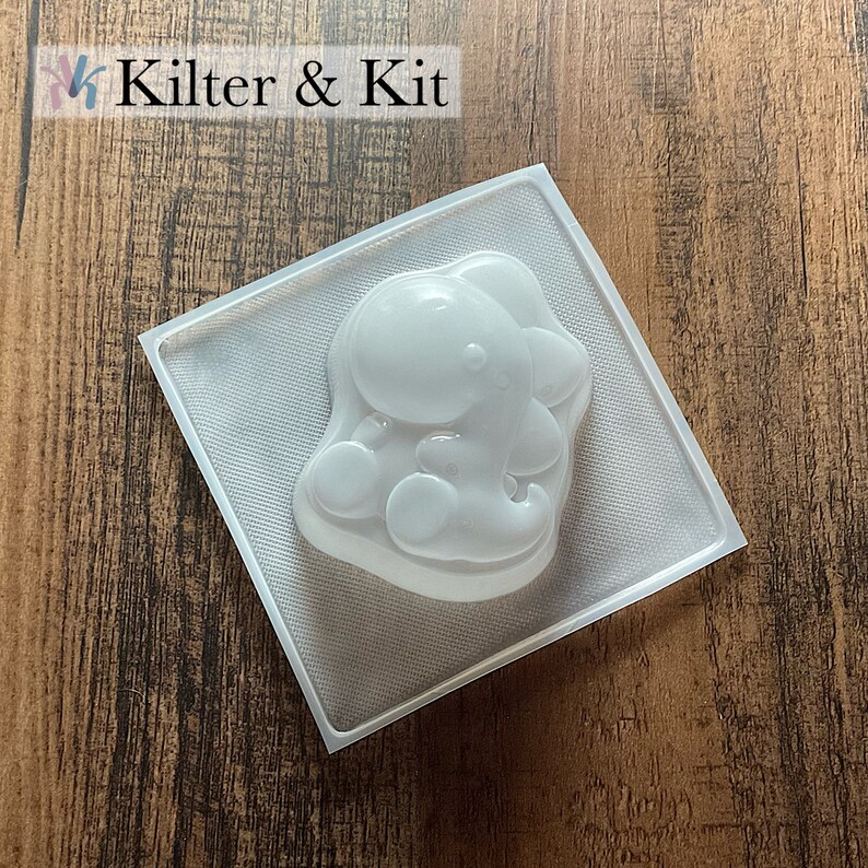 May include: Clear plastic mold for making resin crafts. The mold is shaped like a cartoon dinosaur sitting down with its arms crossed.