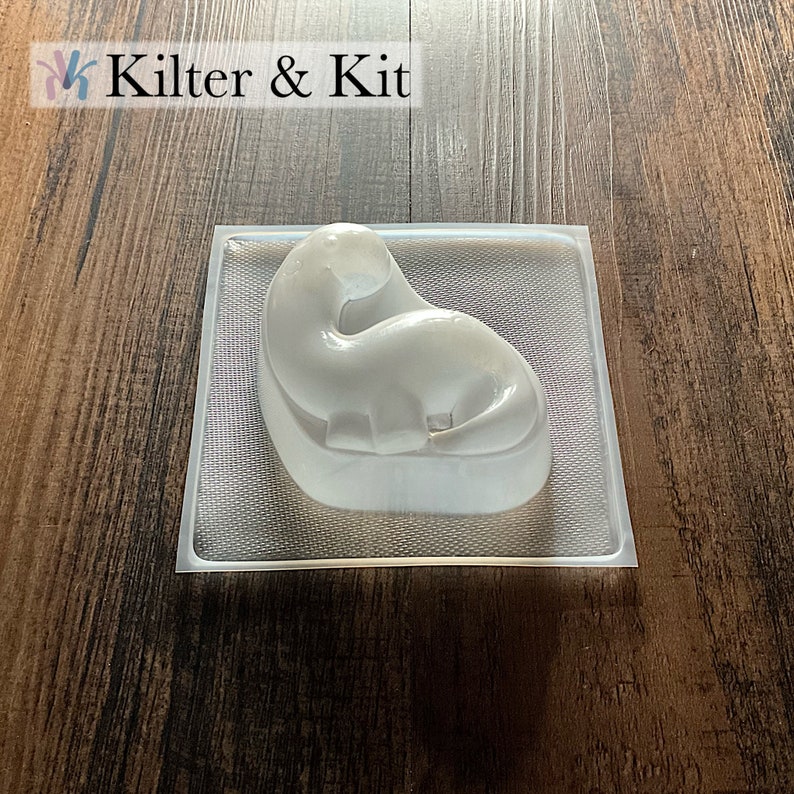 May include: A clear plastic mold of a dinosaur, the dinosaur is curled up in a sleeping position. The mold is on a wooden surface.