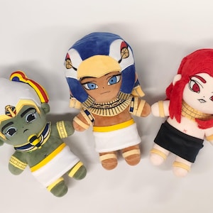 May include: Three plush dolls with Egyptian-inspired designs. One doll has a green face, white headdress, and yellow and red accents. Another has a blue headdress and tan skin. The third doll has red hair and a black skirt.