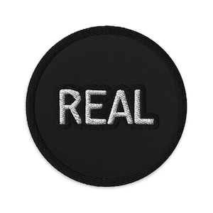 Puede incluir: Un parche circular negro con la palabra "REAL" bordada en blanco en el centro.