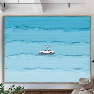 May include: Large framed abstract art piece with a light blue textured background. A white sports car with a figure on top is centered. The artwork is displayed in a bright, modern living room setting.