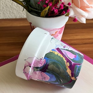 May include: Two white ceramic flower pots with a marbled design. The larger pot has a green, blue, pink, and gold marbled design. The smaller pot has a pink and white marbled design.