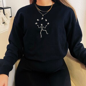 May include: Black sweatshirt with a white skeleton juggling skulls. The design is centered on the front of the long-sleeved crew neck sweatshirt. The skeleton is shown with its arms and legs outstretched, with six skulls floating above it.