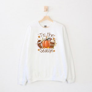 May include: White sweatshirt with a fall-themed graphic. The design includes a football, pumpkins, a coffee cup, and fall leaves. The text "Tis the Season" is also featured in the design. The sweatshirt is hanging on a wooden hanger.