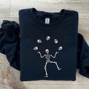 May include: A navy blue sweatshirt featuring a white skeleton juggling skulls. The skeleton is in the center of the sweatshirt, with the skulls floating above it. The sweatshirt is made of a soft material.