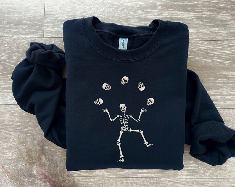 May include: A navy blue sweatshirt featuring a white skeleton juggling skulls. The skeleton is in the center of the sweatshirt, with the skulls floating above it. The sweatshirt is made of a soft material.