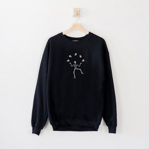 May include: Black sweatshirt featuring a white illustration of a skeleton juggling skulls. The garment is suspended on a wooden hanger against a white wall. The design is centrally placed on the front.