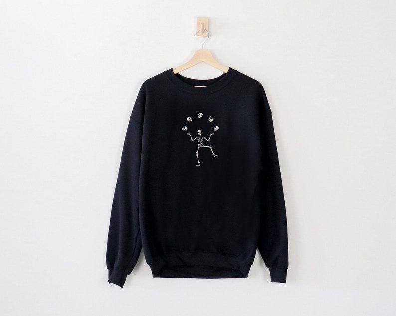 May include: Black sweatshirt featuring a white illustration of a skeleton juggling skulls. The garment is suspended on a wooden hanger against a white wall. The design is centrally placed on the front.