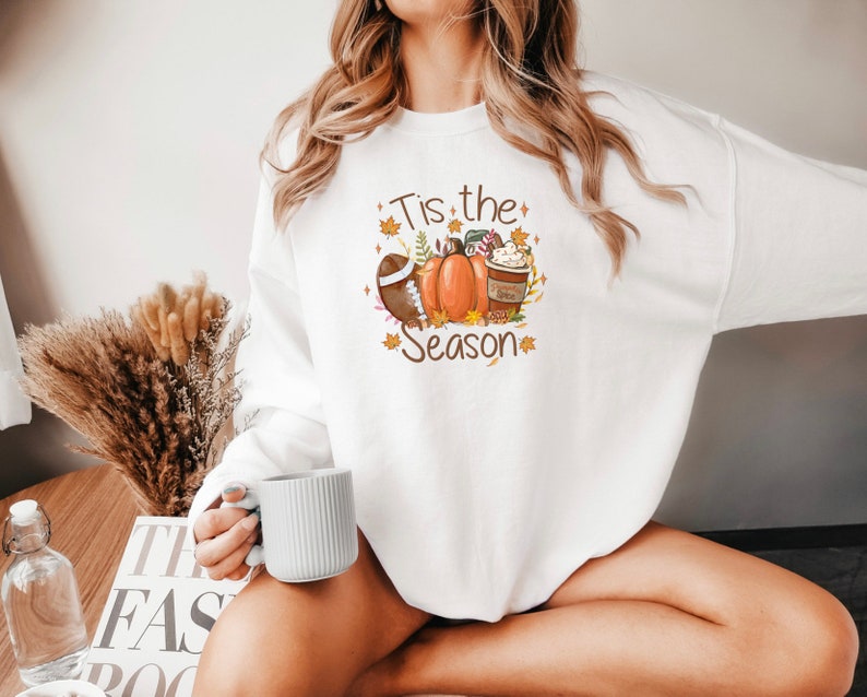May include: White sweatshirt with a fall-themed graphic. The design includes a football, pumpkins, a coffee cup, and the text "Tis the Season" in brown. Autumn leaves and decorative elements surround the central images.