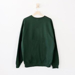 May include: A dark green sweatshirt hangs on a wooden hanger against a white wall. The crew neck sweatshirt is made of a soft material and has long sleeves. The garment is displayed from the back.